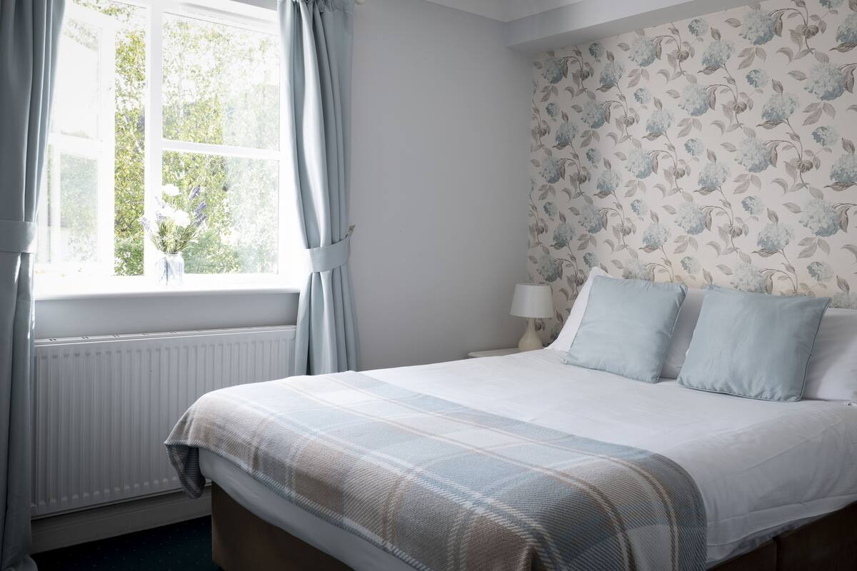 Een slaapkamer met een bed, een raam met gordijnen, een radiator en bloemenbehang.