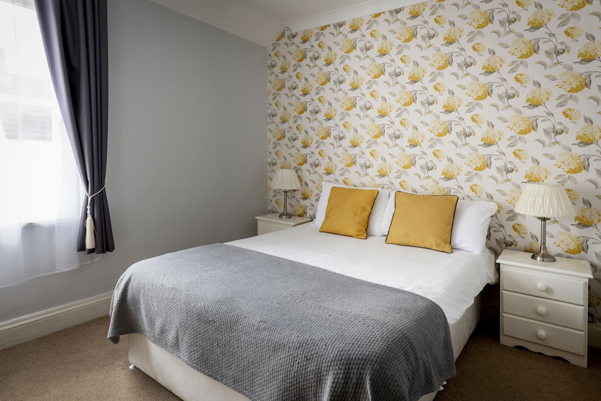 Een slaapkamer met een bed, twee nachtkastjes, een lamp op elk en een raam met gordijnen, plus behang met bloemen.