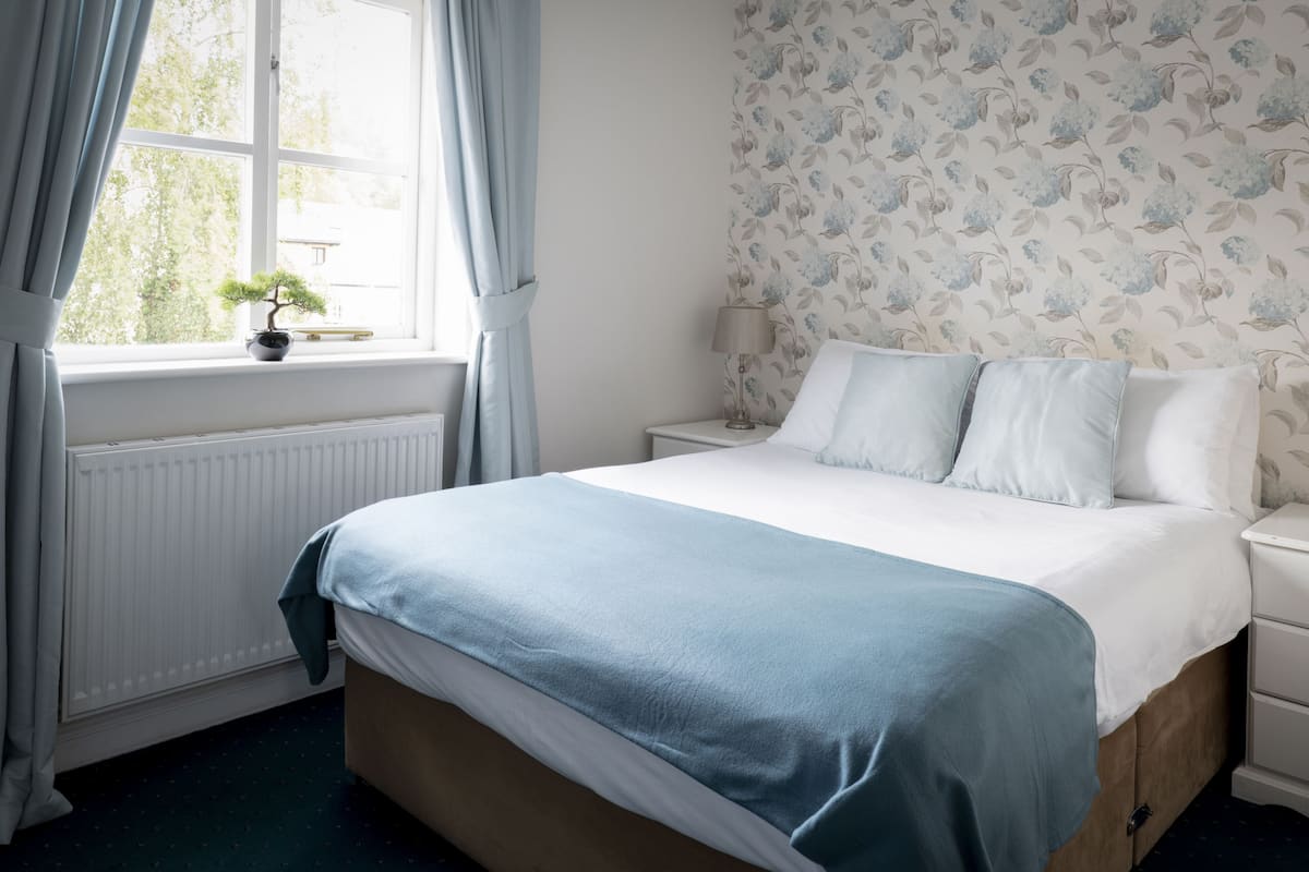 Een slaapkamer met een bed, nachtkastje, lamp, raam met gordijnen en een radiator.