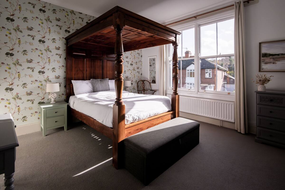 Een hemelbed met houten frame, een nachtkastje met lamp, een bankje en een dressoir met spiegel.