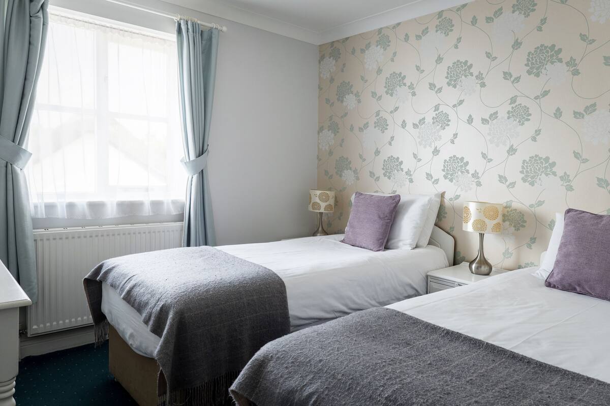 Een hotelkamer met twee bedden, een raam met gordijnen, een radiator en bloemenbehang.