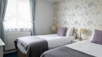 A hotel room with two beds, a window with curtains, a radiator, and floral wallpaper.