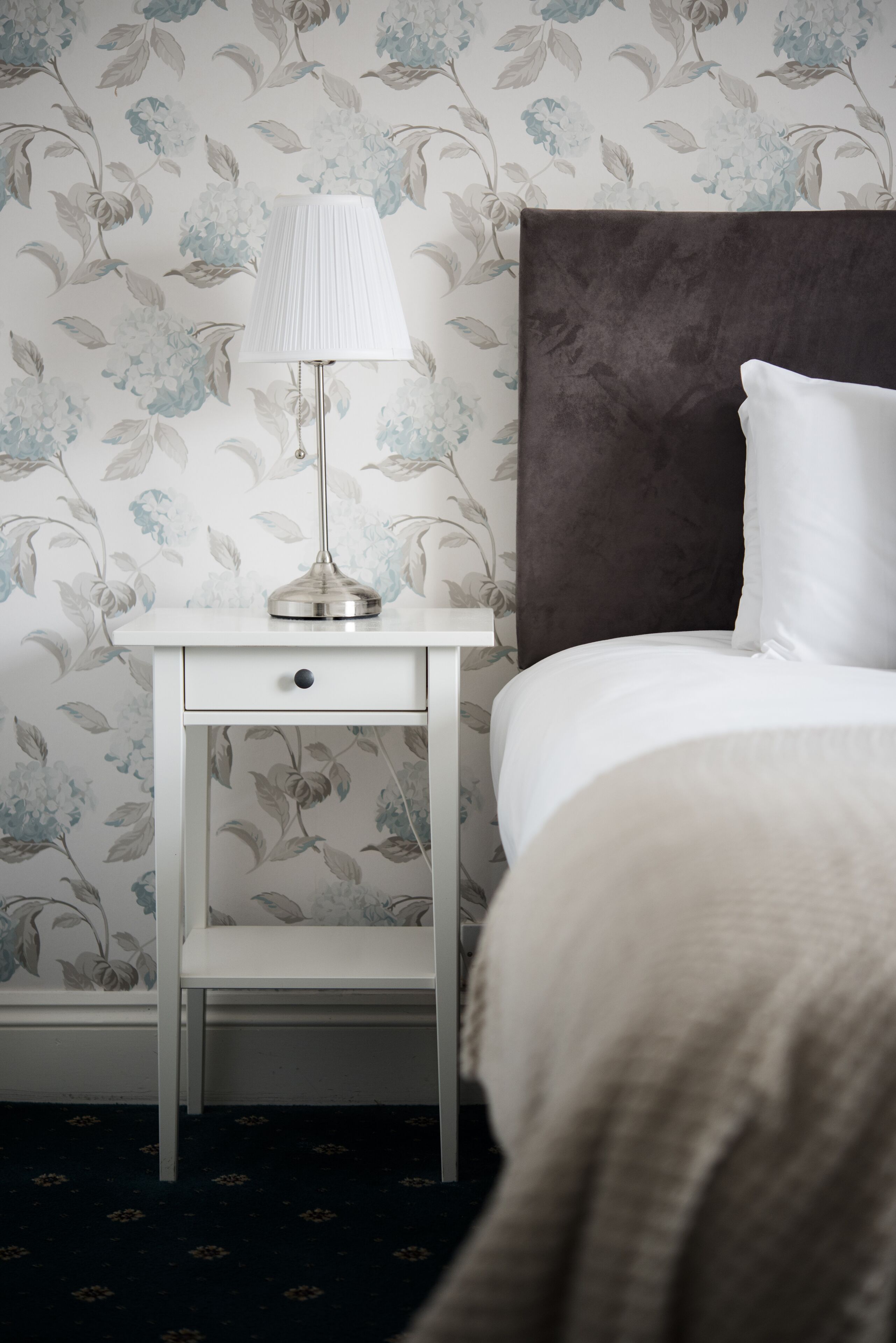 A neatly made bed with a white pillow and a white bedside table with a lamp.