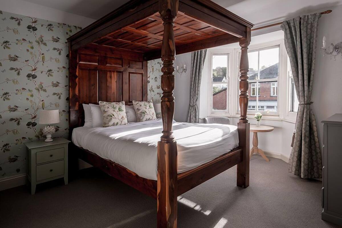Een hemelbed met houten frame, een nachtkastje in het groen met een lamp, bloemenbehang en een uitzicht op gebouwen door een raam met gordijnen.