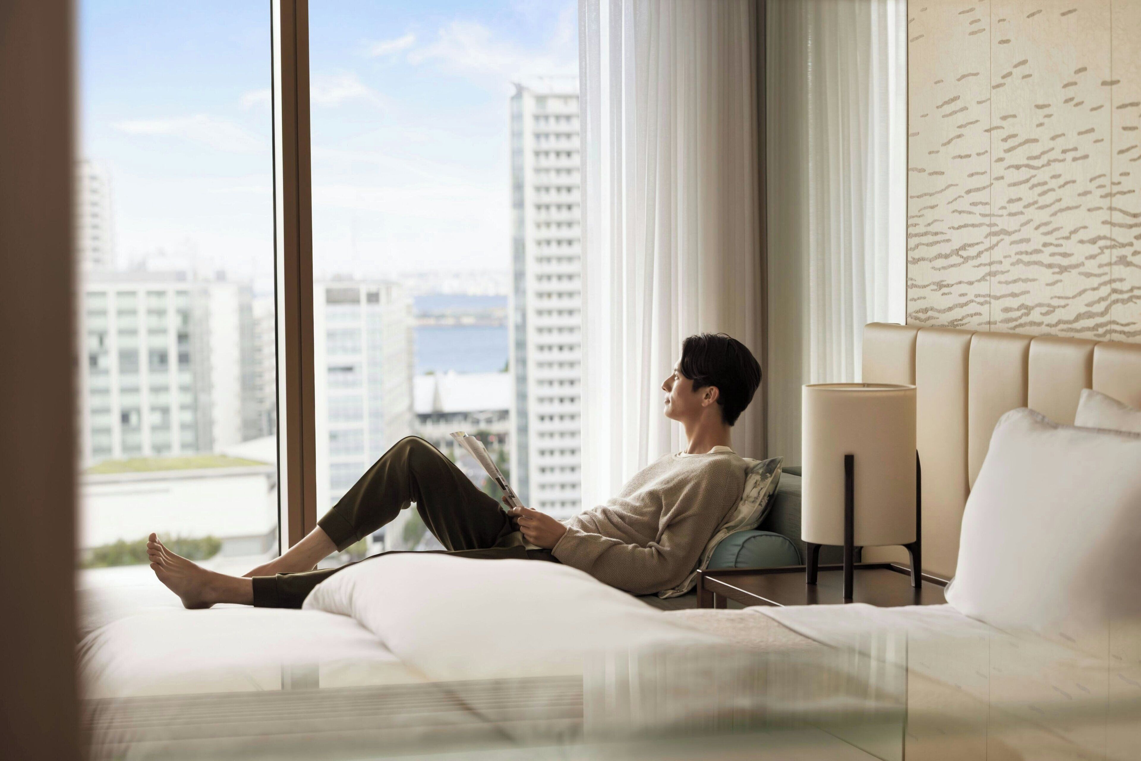 Iemand zit op een bed in een moderne hotelkamer met uitzicht op de stad, een groot raam en een wandlamp.