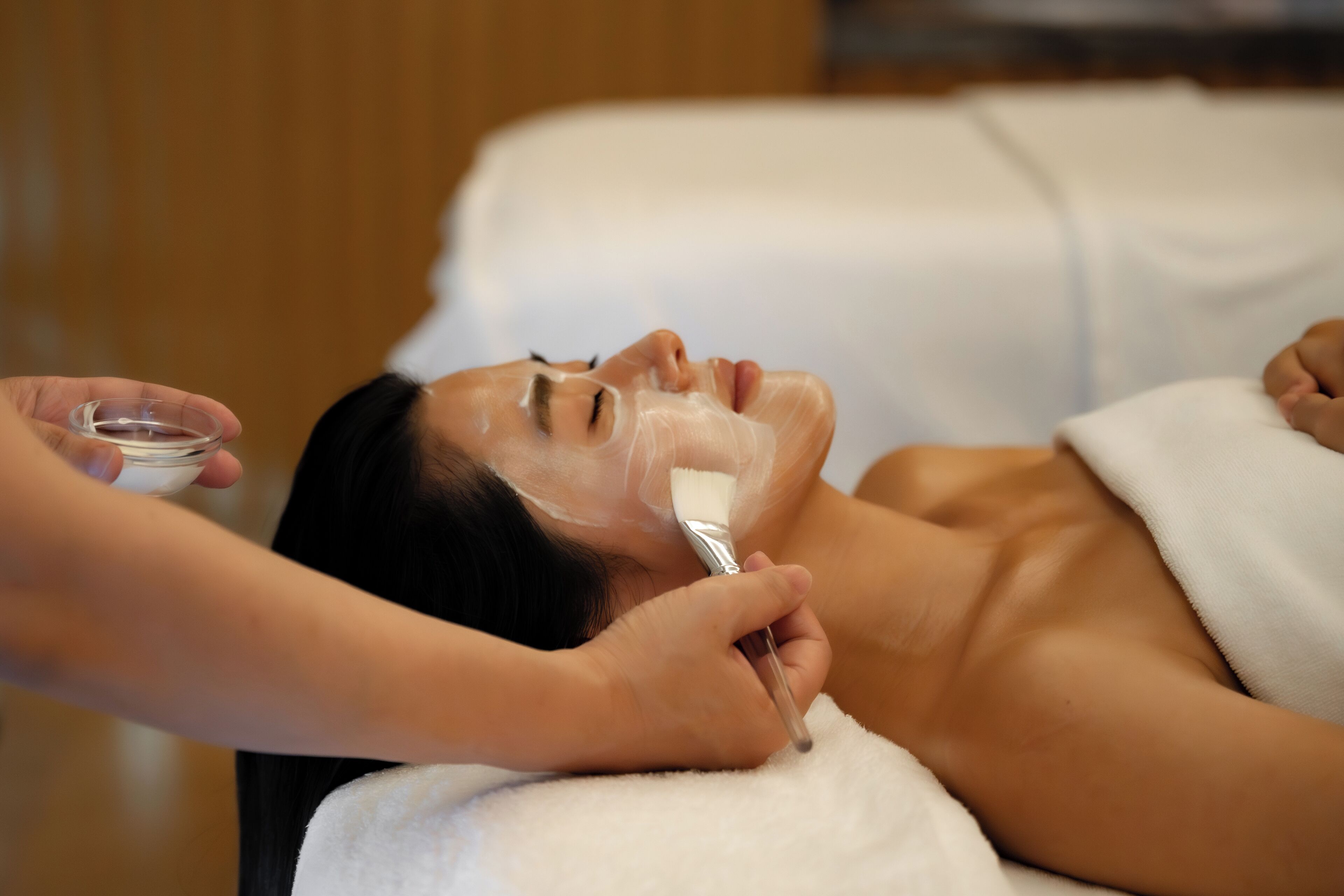 A person receiving a facial treatment in a room with a white towel and wooden paneling.