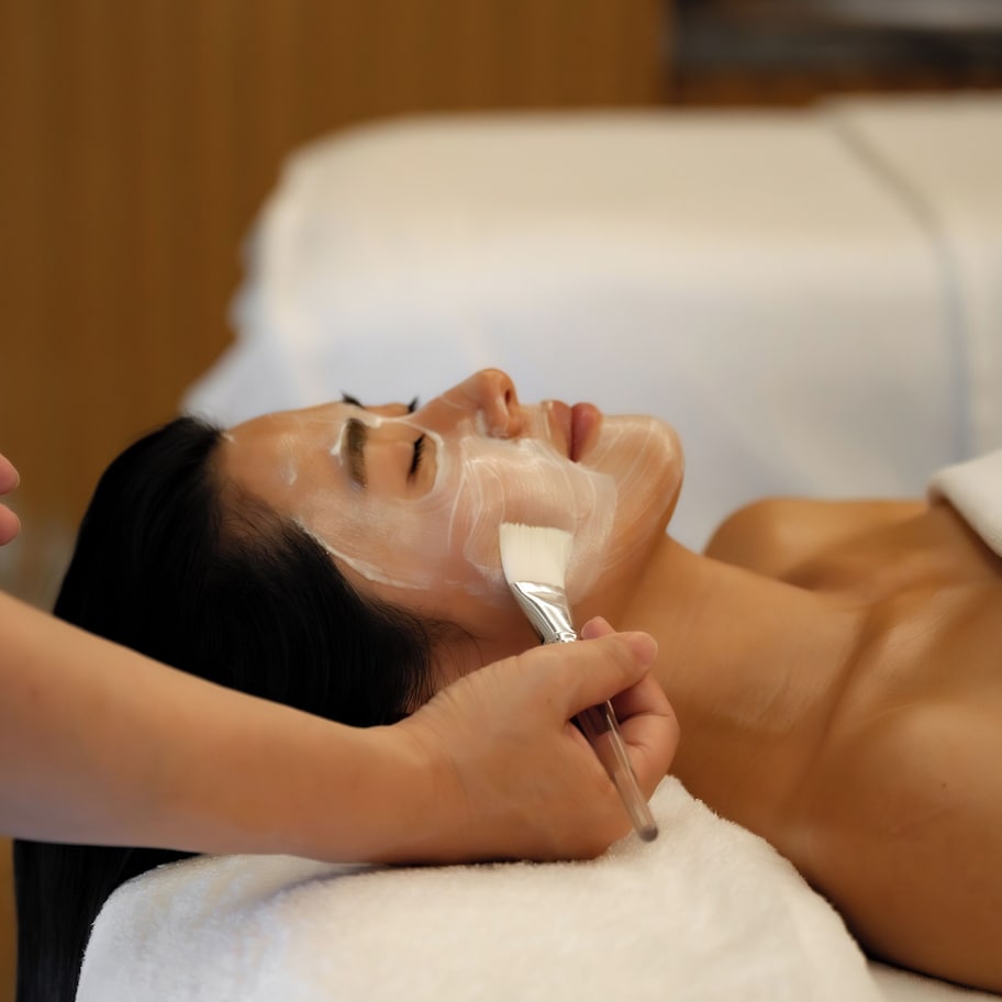 A person receiving a facial treatment in a room with a white towel and wooden paneling.