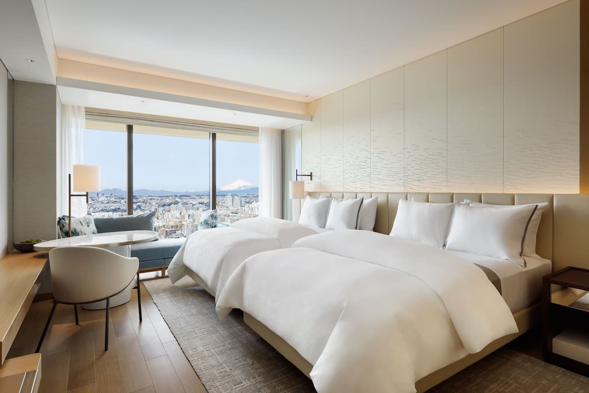 Habitació d'hotel moderna amb un llit gran, una taula de treball i una cadira. Es veu un paisatge urbà i una muntanya al fons.