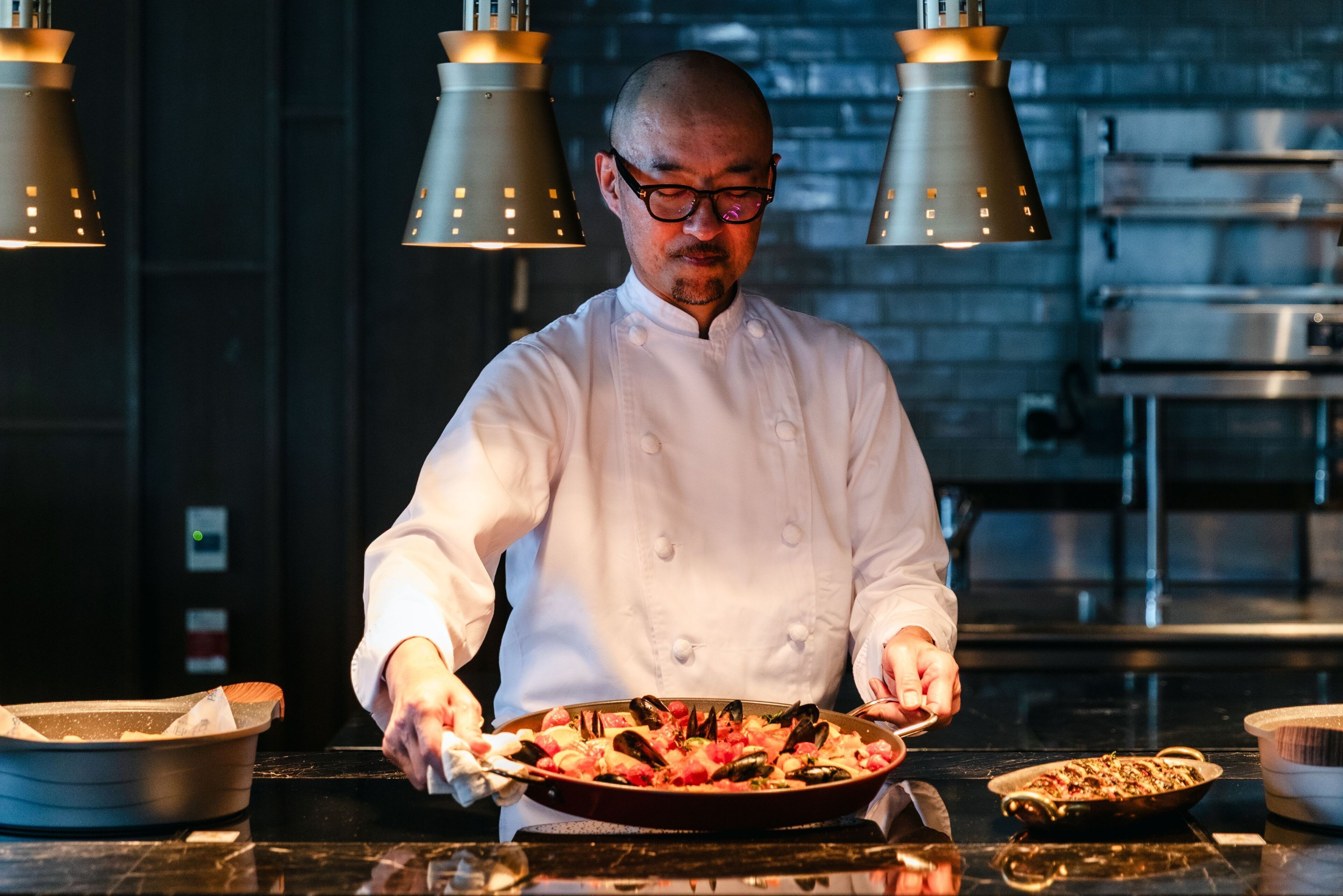 A chef in a white uniform preparing a dish in a kitchen with modern lighting and stainless steel appliances.