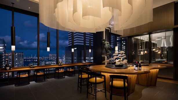 Un moderno bar con vistas panorámicas a la ciudad, una lámpara colgante grande y una cocina elegante visible a través de cristales.