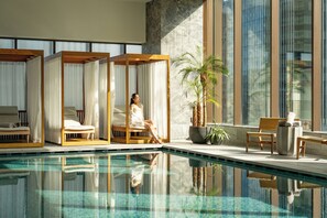 A poolside area with a clear pool, wooden seating, and a large window with a view.