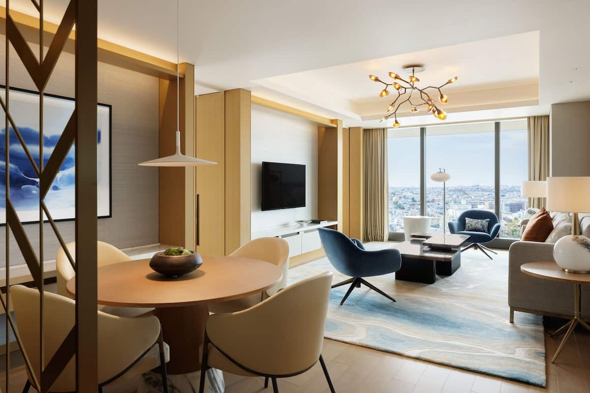Habitación de hotel moderna con zona de comedor, asientos, televisor y ventana grande con vista a la ciudad.