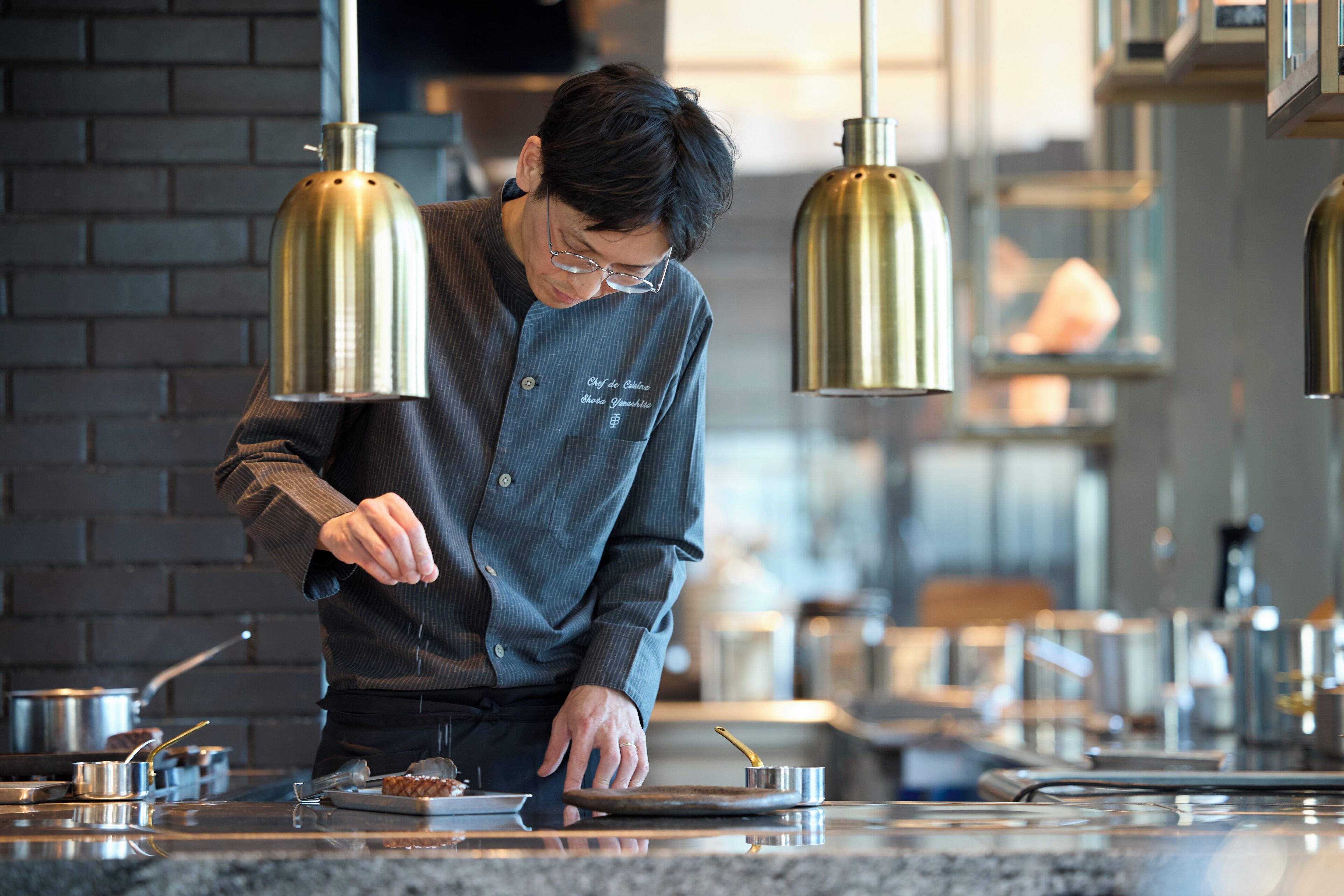 A person cooking in a modern kitchen with brick walls and pendant lighting.