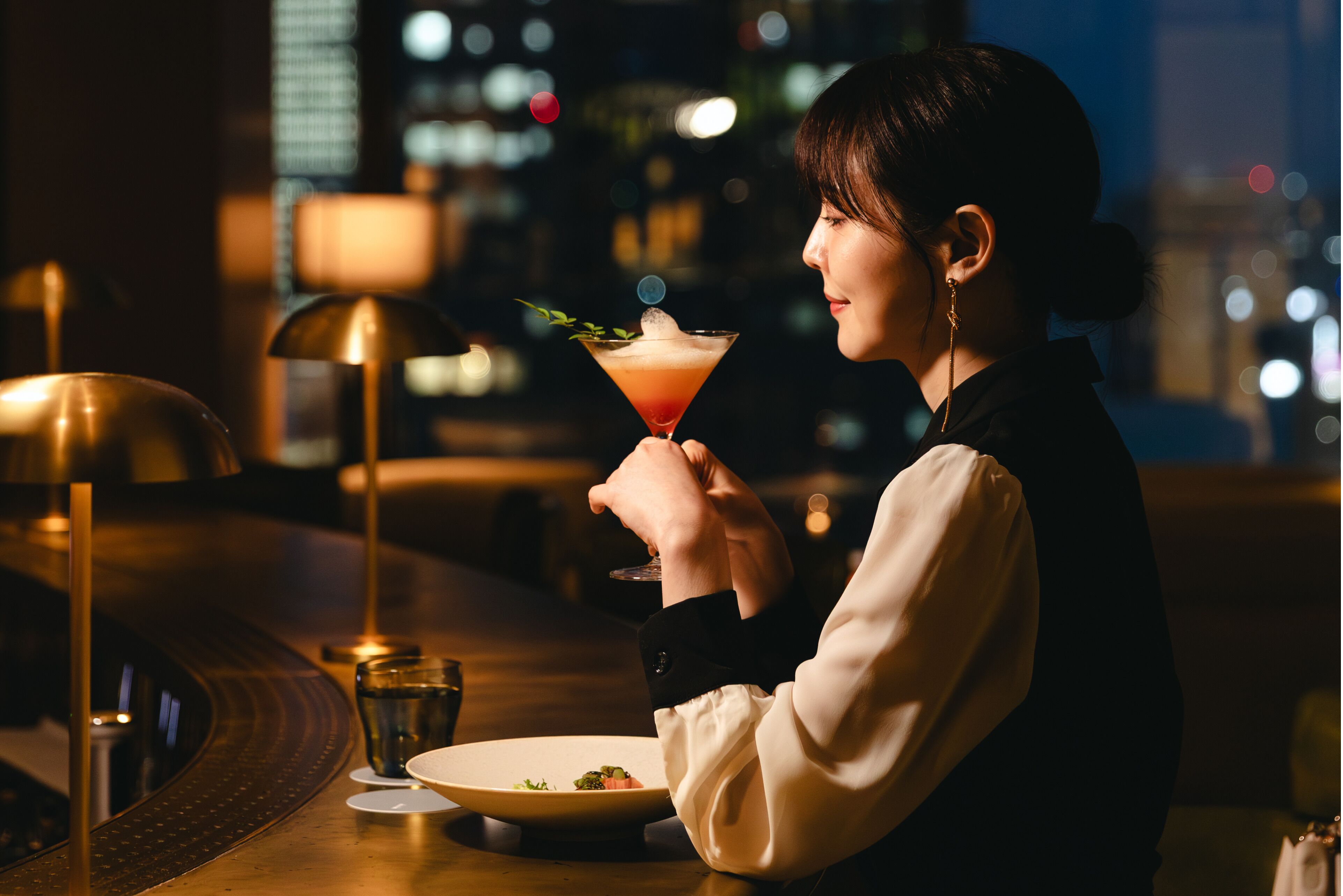 A person holding a cocktail with a garnish, seated at a table with a plate of food, in a dimly lit indoor setting with a cityscape view.