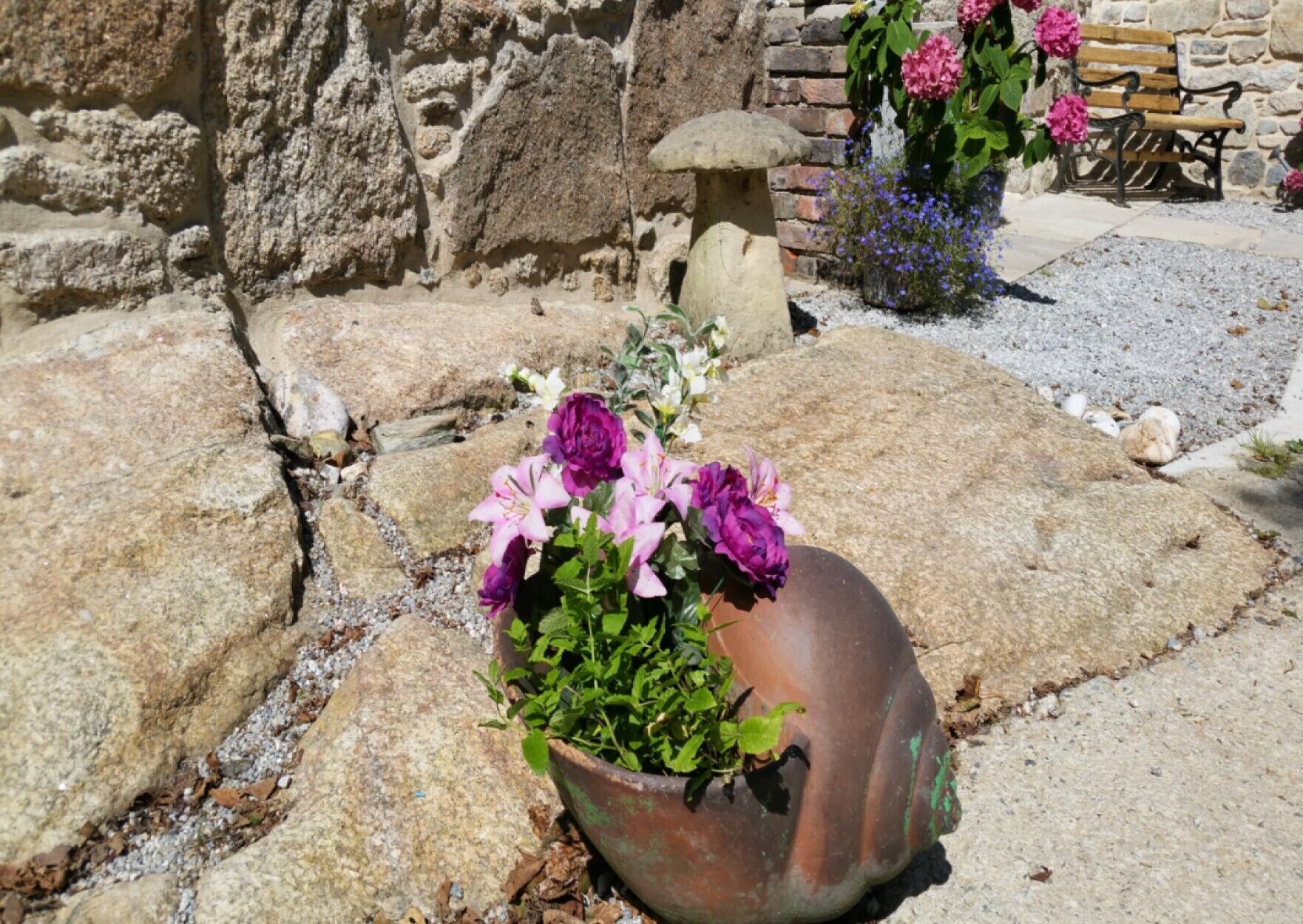 A stone pathway with a flower pot containing pink and purple flowers, a stone bench, and a stone wall.