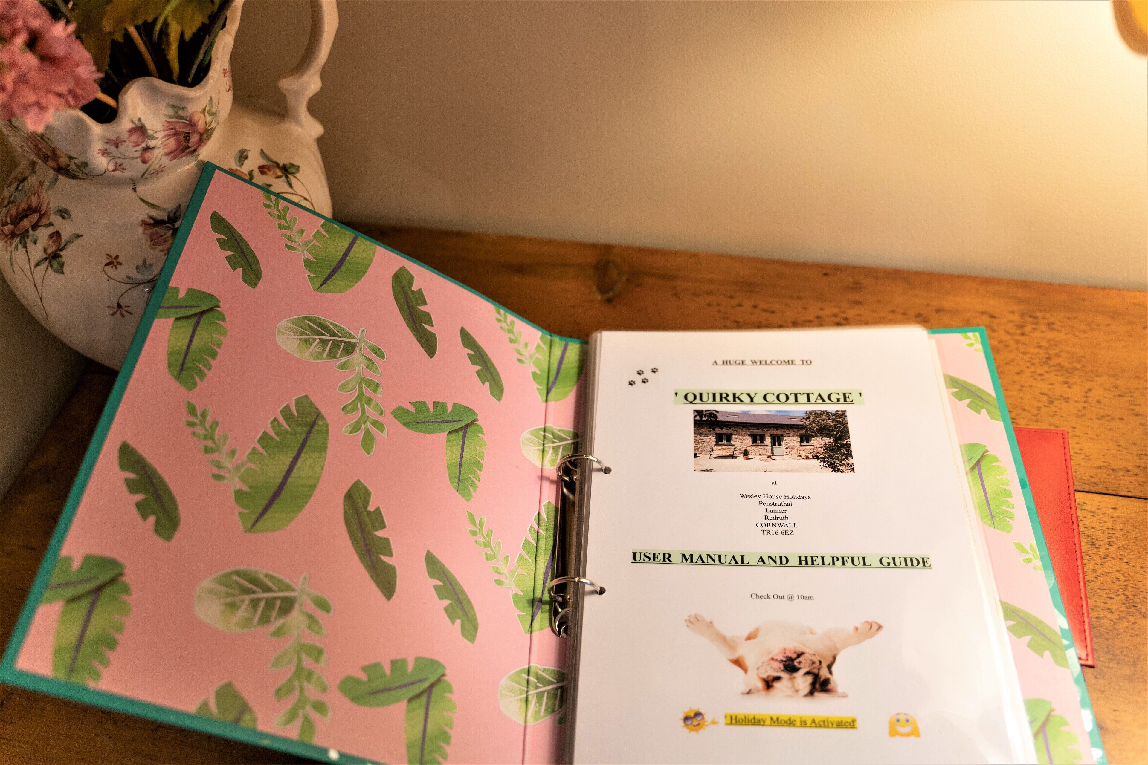 A binder with a pink cover featuring green leaf patterns, open to a page titled 'QUIRKY COTTAGE' with a photo of a house and text about holiday mode.
