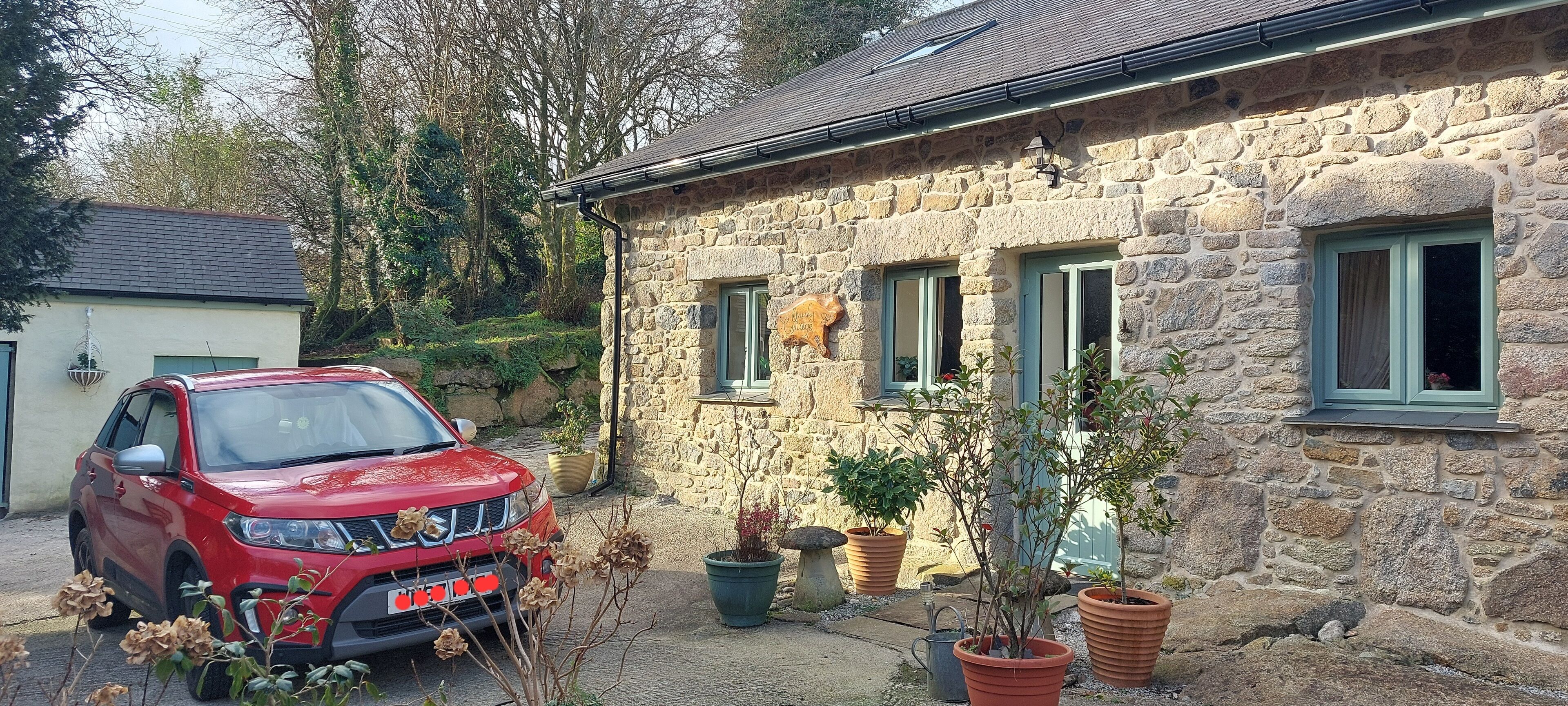 A stone house with a red car parked in front, potted plants, and a small garden area.