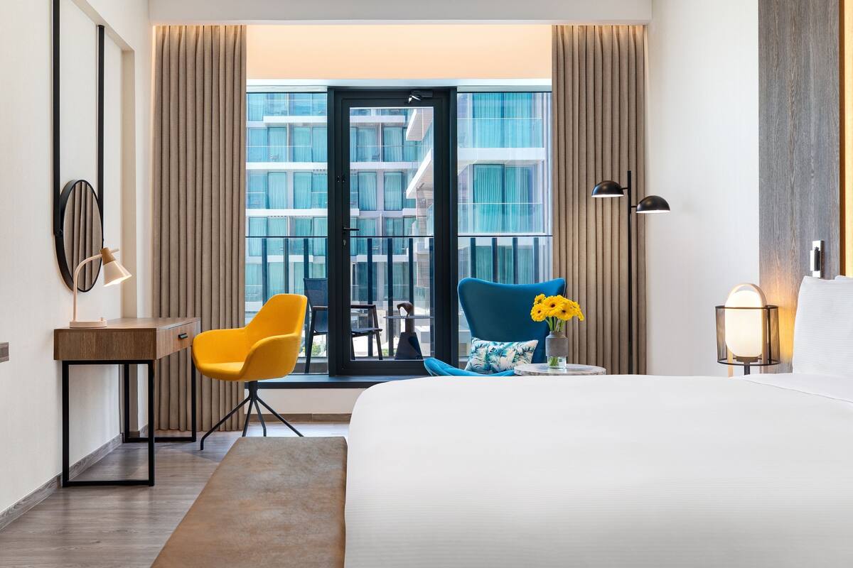 Une chambre d’hôtel avec un lit, un bureau avec une lampe, une chaise, une petite table avec un vase de fleurs et une vue sur les bâtiments par la fenêtre.