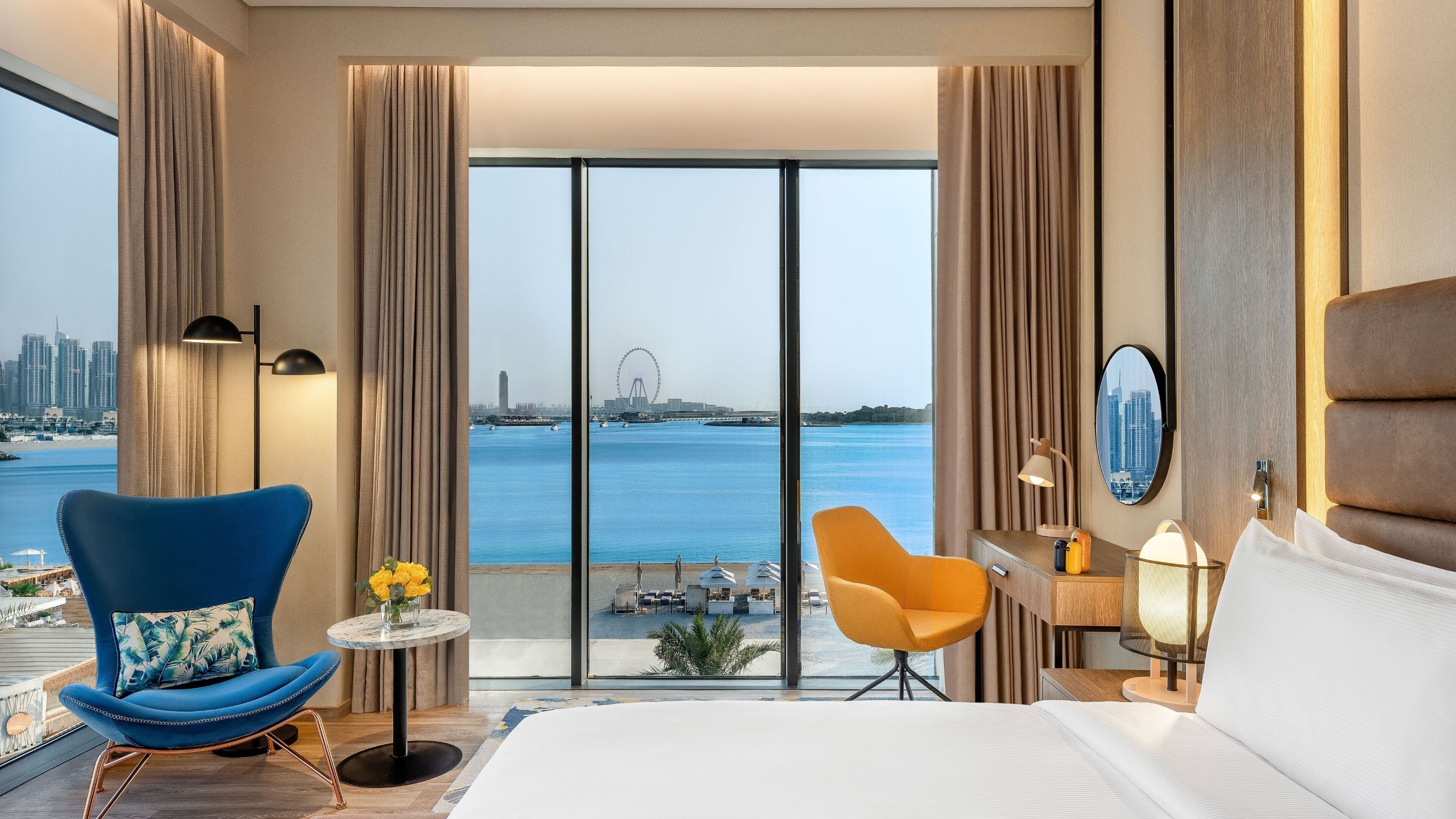 Habitación de hotel con una cama grande, un sillón azul, una mesita con un florero y vistas a un horizonte urbano y al agua.