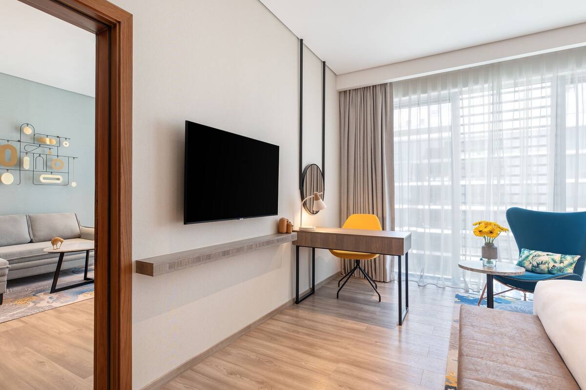 Une chambre d’hôtel moderne dotée d’un téléviseur à écran plat fixé au mur, d’un canapé, d’un bureau avec une chaise et d’une grande fenêtre avec des rideaux légers.