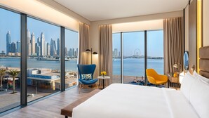 Camera d'albergo moderna con un letto grande, una poltrona blu, una sedia gialla e vista sullo skyline cittadino e sul mare.