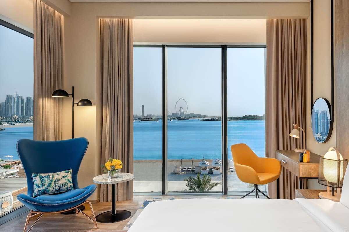 Une chambre d’hôtel avec un grand lit, un fauteuil bleu, une petite table ronde ornée d’un vase de fleurs, et une vue sur la mer et le panorama de la ville.