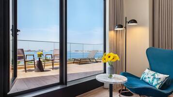Una habitación moderna con vista al mar, una silla azul con un cojín estampado y un jarrón con flores amarillas.