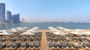 A beach with rows of sun loungers and umbrellas, a clear view of the sea, and a city skyline in the background. - voco Dubai the Palm by IHG (Dubai)