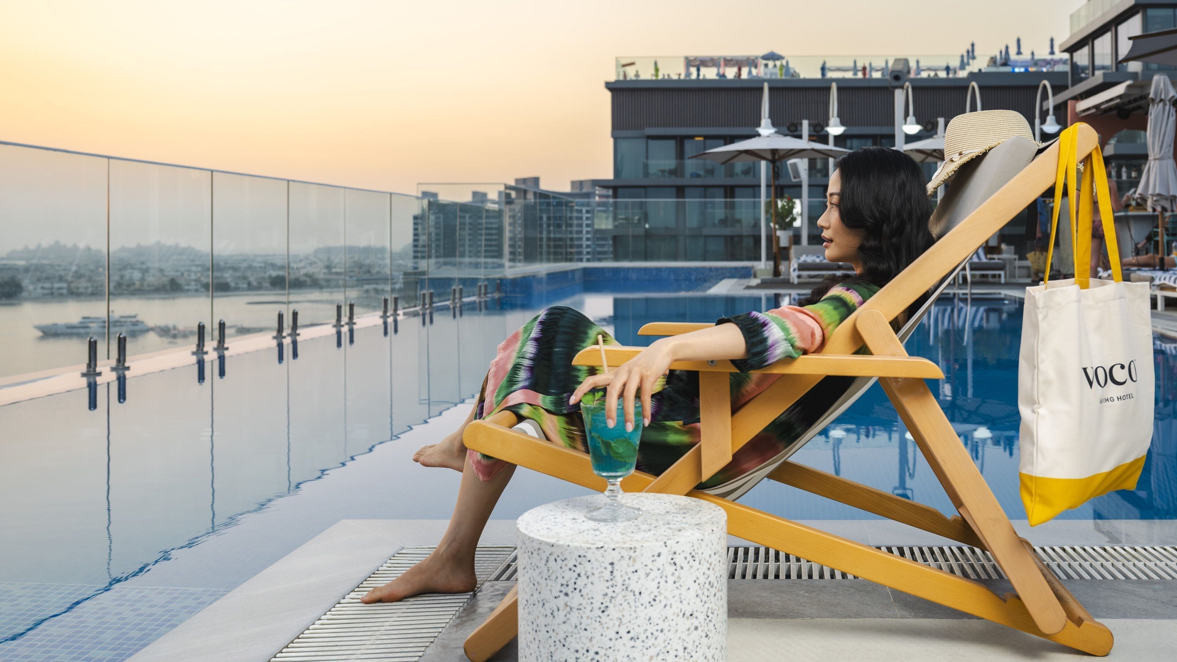 Eine Person entspannt auf einer Liege am Pool mit Blick auf die Stadt, einer weißen Tragetasche und einem Getränk.