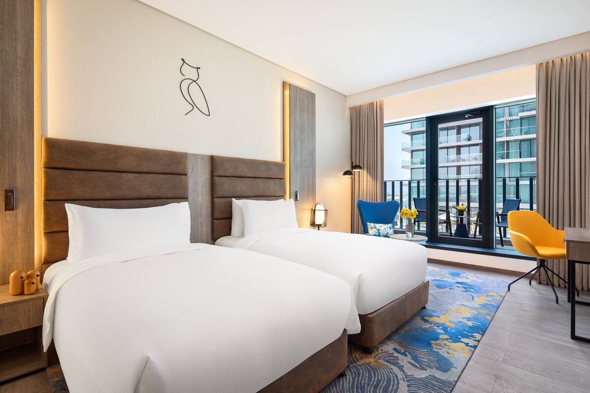 Une chambre d’hôtel avec deux lits, un bureau, une chaise, une lampe et une vue sur un immeuble, visible à travers une porte vitrée.