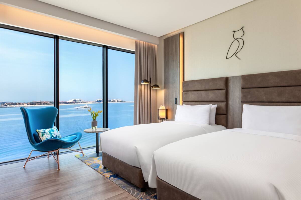 Une chambre d’hôtel avec deux lits, une vue sur la mer, une chaise bleue et une petite table sur laquelle est posé un vase de fleurs.