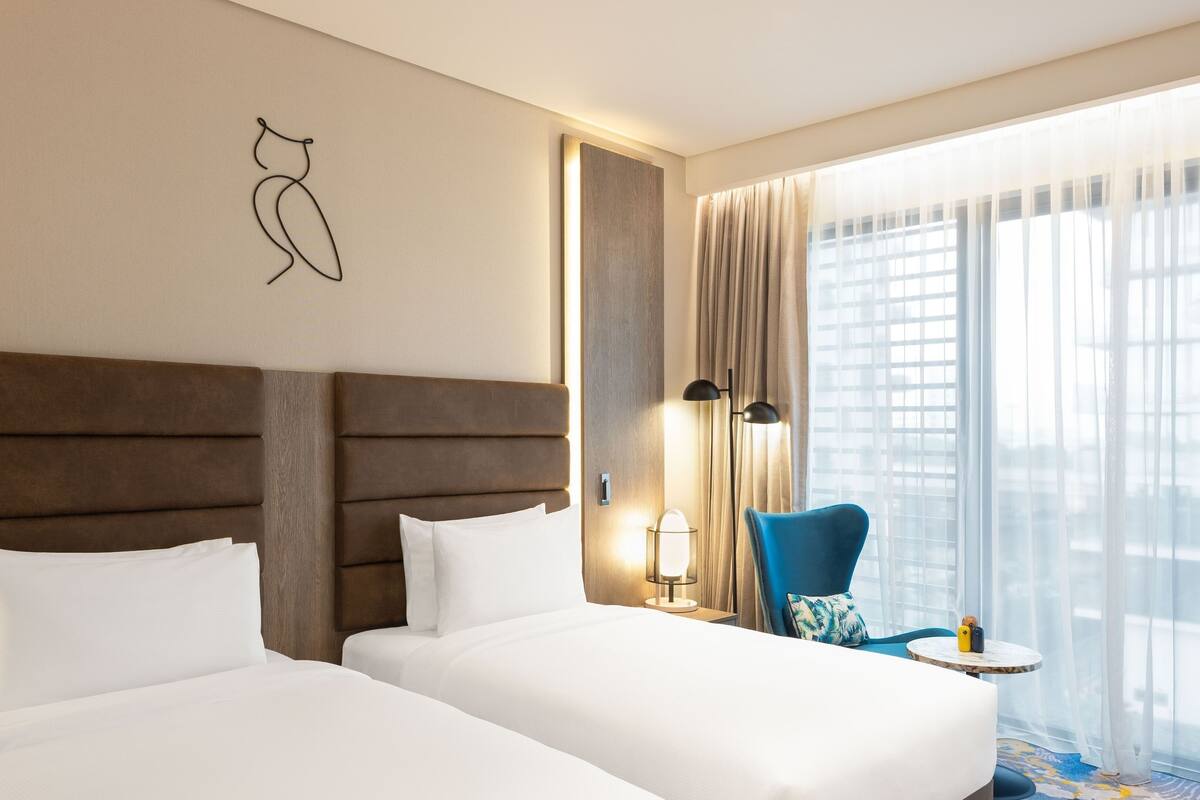 Une chambre d’hôtel avec deux lits, une grande fenêtre avec des rideaux, une lampe sur pied et une chaise bleue.