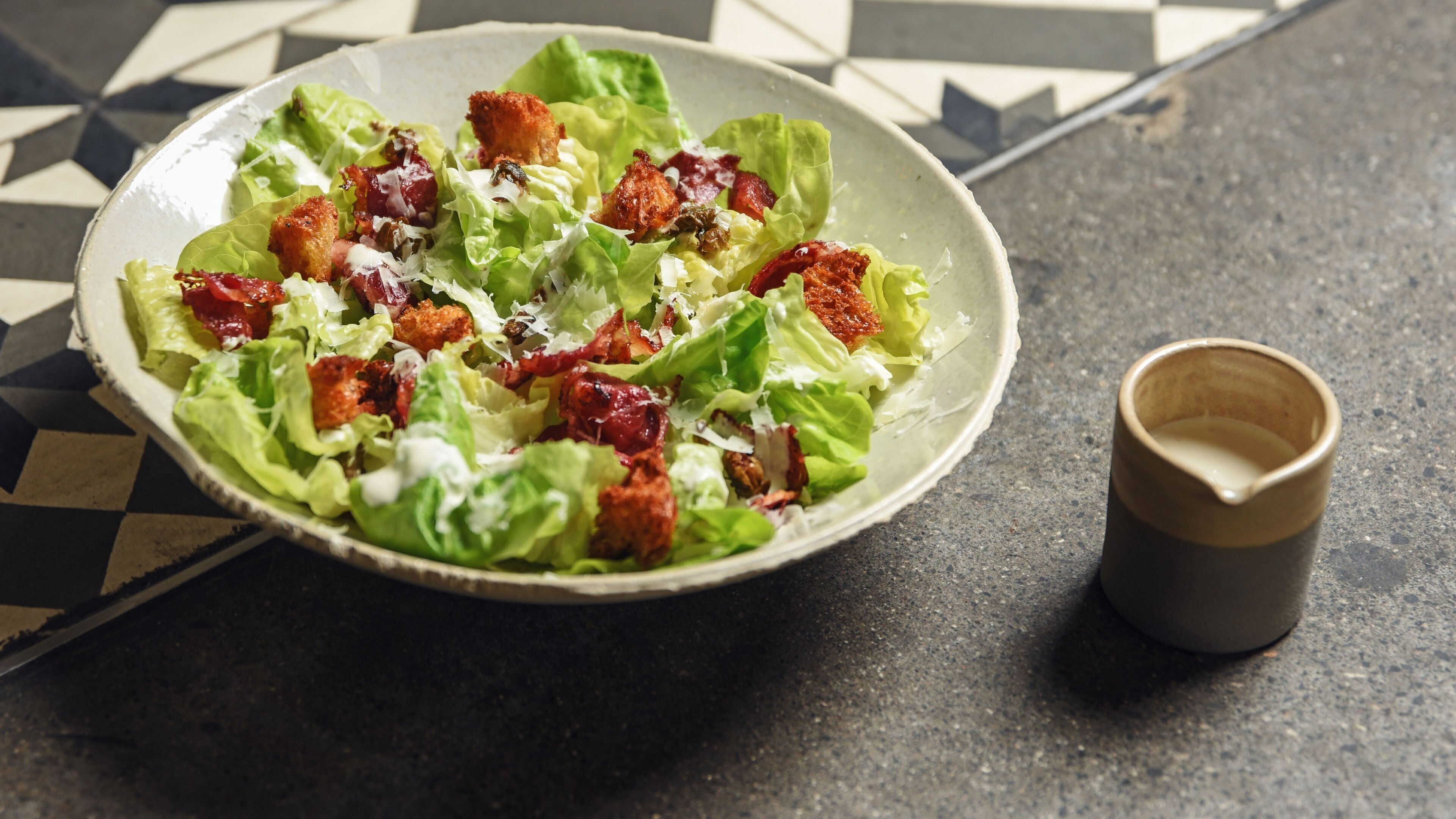 Een saladekom met croutons en spek, op een betegelde vloer.