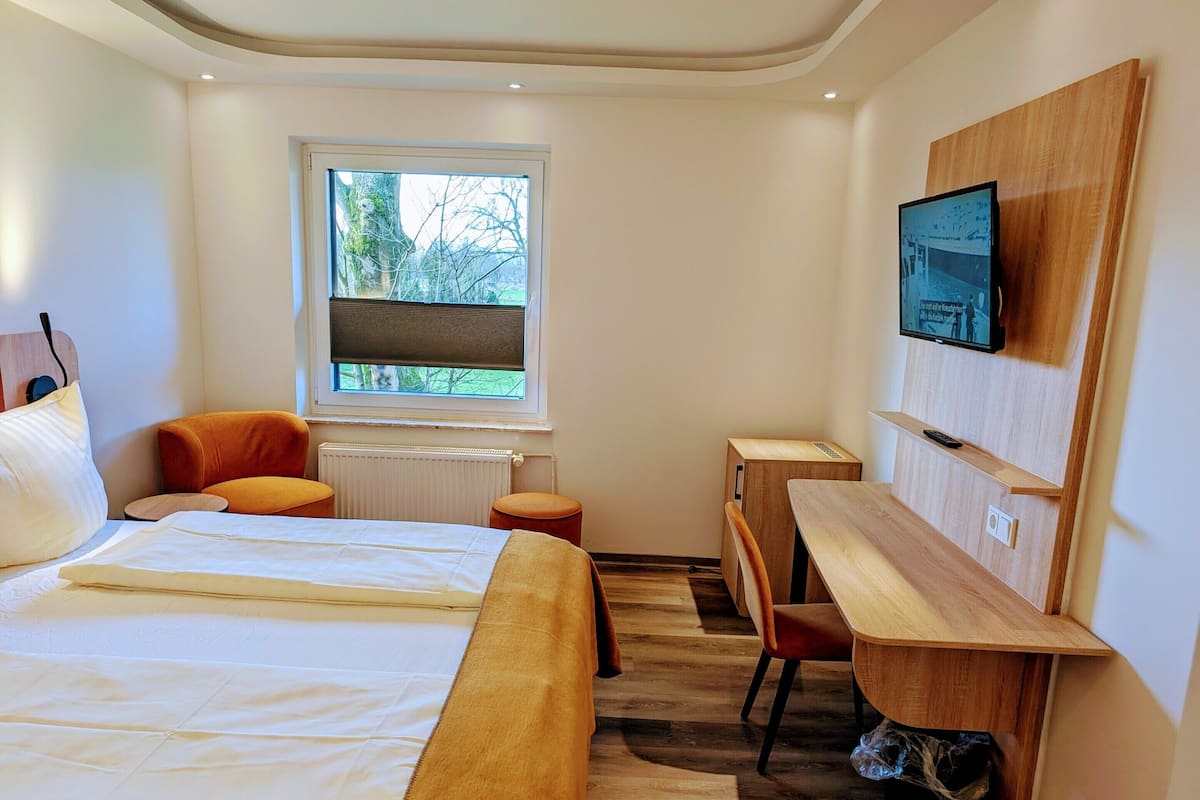 A modern hotel room with a large bed, a desk, a chair, a TV, and a window with a view of trees.