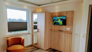 A modern room with a wooden cabinet, a television mounted on the wall, a window with a view of trees, and a brown leather chair. - Hotel Tausendschön (Sudbrookmerland)