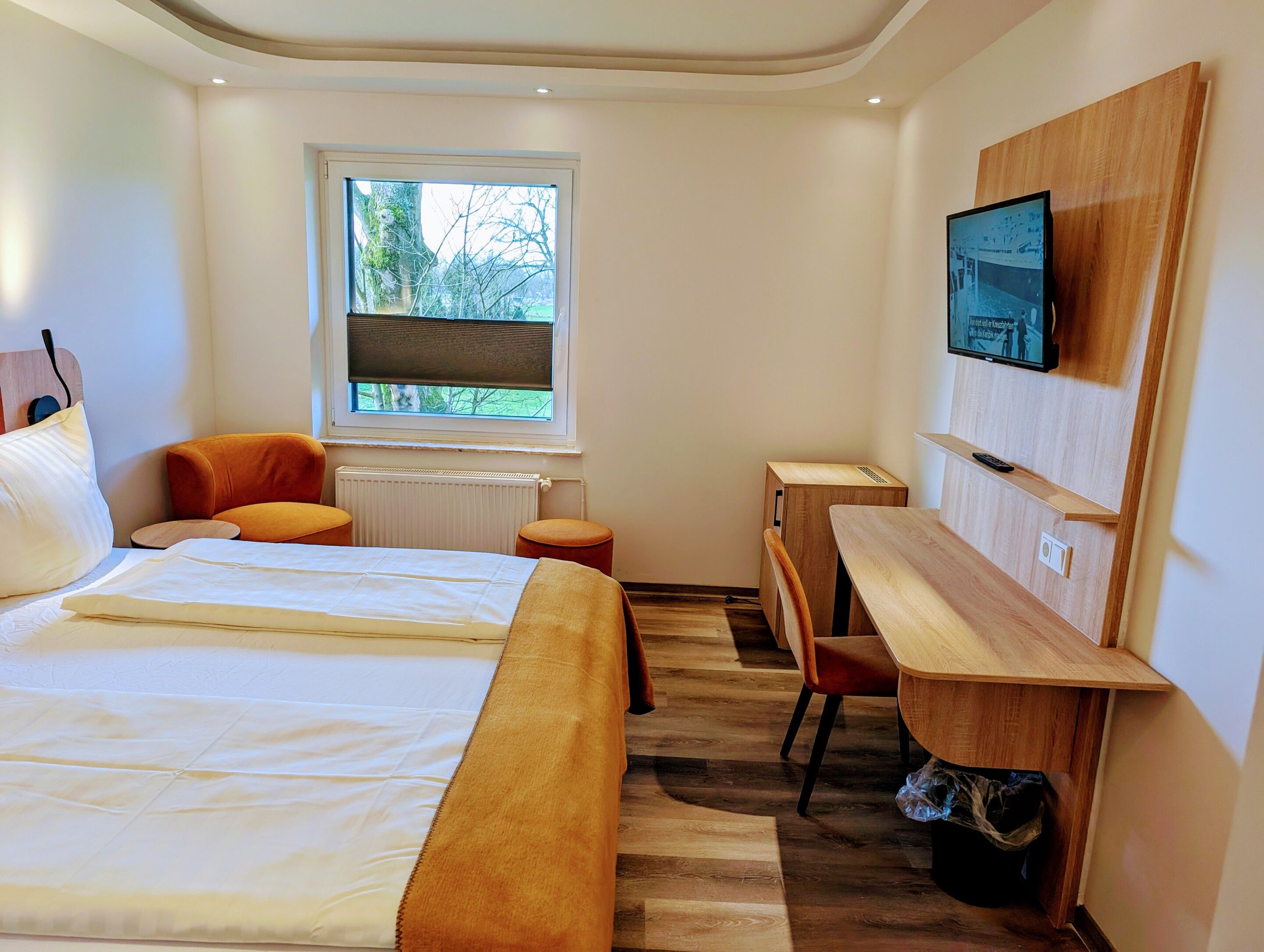 A modern hotel room with a large bed, a desk, a chair, a TV, and a window with a view of trees.