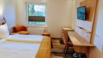 A modern hotel room with a large bed, a desk, a chair, a TV, and a window with a view of trees.
