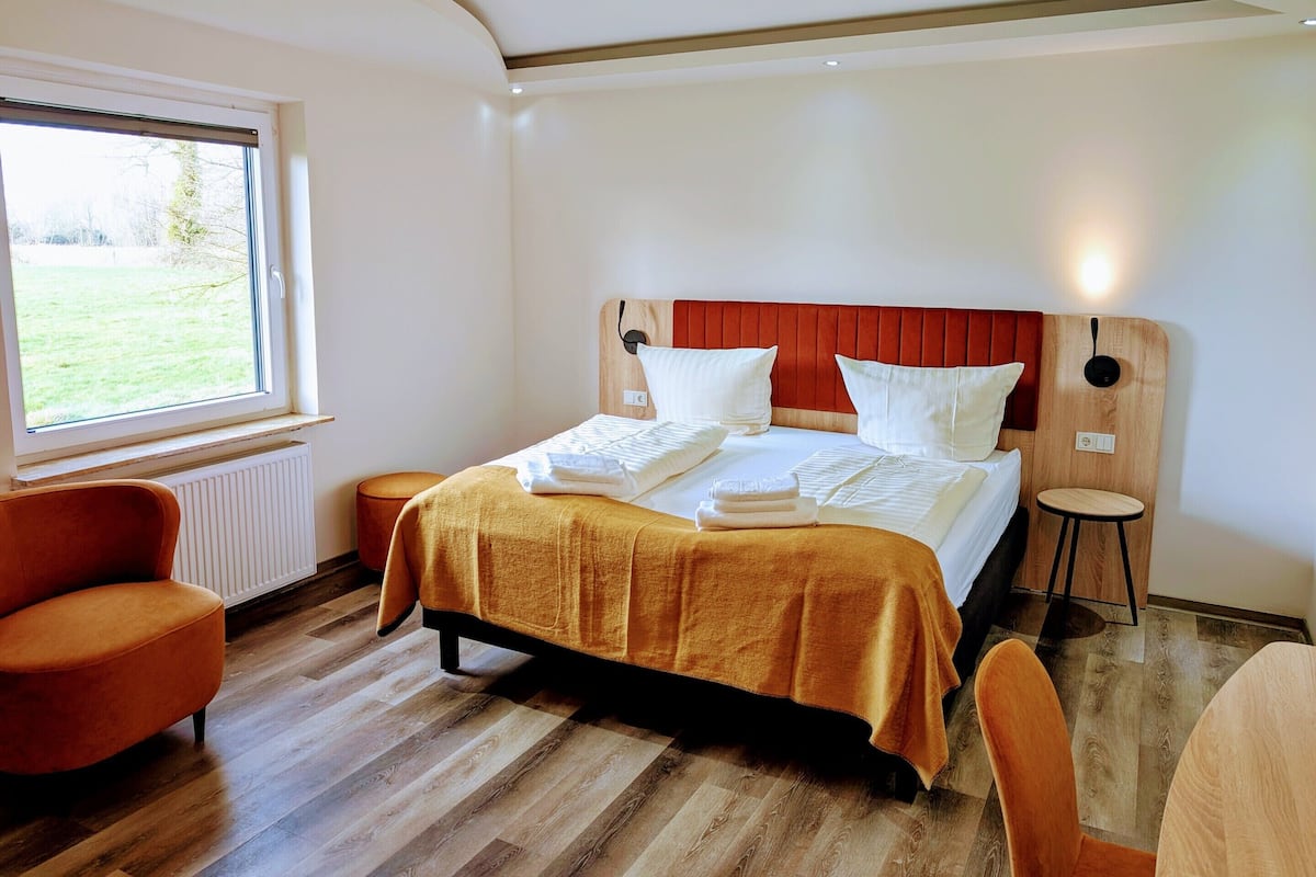A hotel room with a bed, two chairs, a small table, a window with a view of a grassy area, and a wooden headboard.