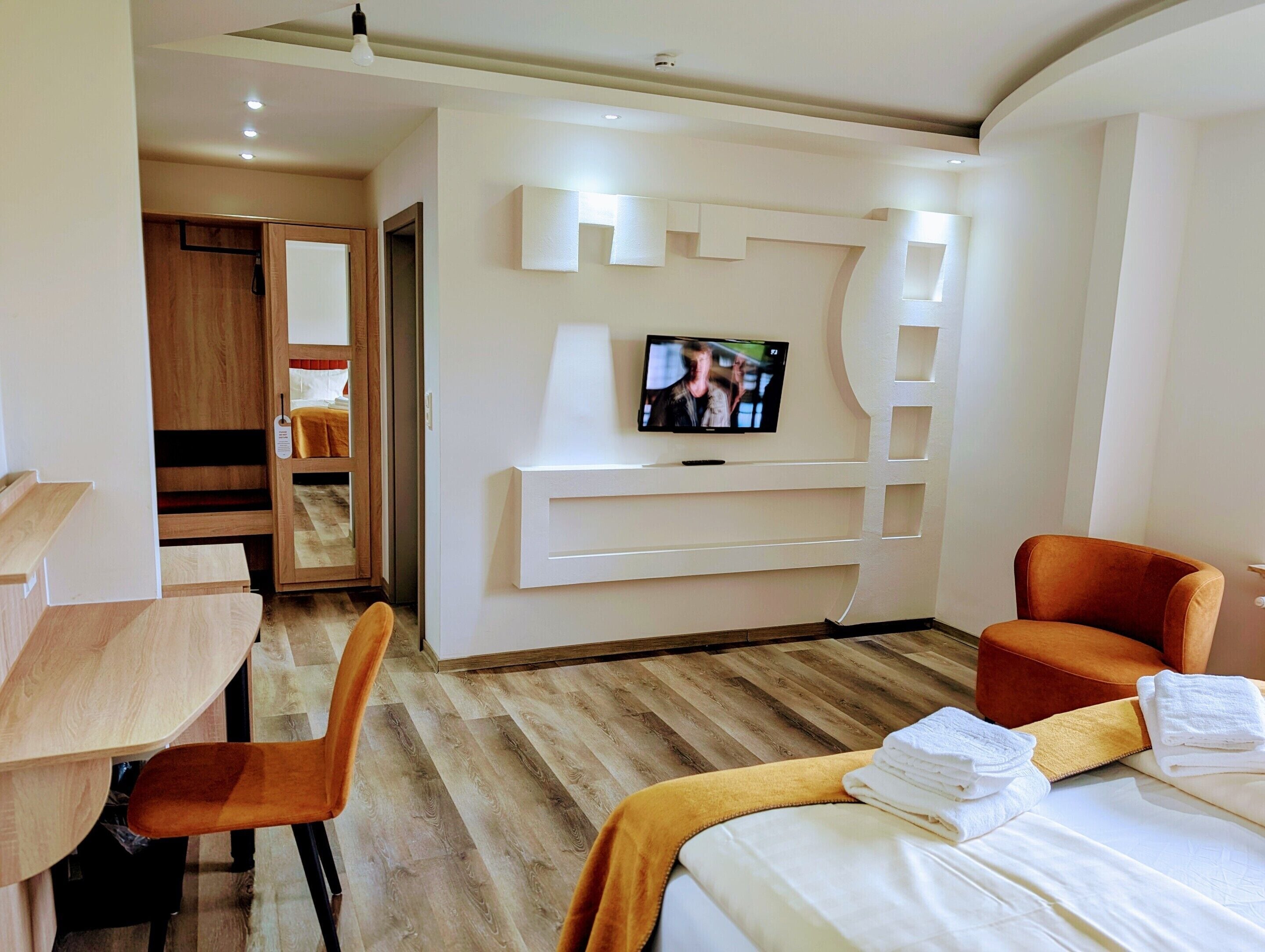 Habitación de hotel con cama, escritorio, silla, televisor montado en la pared y armario de madera.