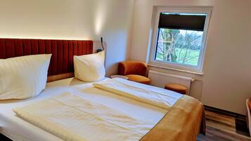 Un lit bien fait, avec des oreillers et une couverture, une fenêtre donnant sur des arbres et une petite chaise orange.