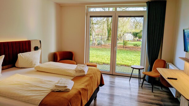 A hotel room with a bed, a desk, a chair, and a large sliding glass door leading to a garden. - Hotel Tausendschön (Sudbrookmerland)