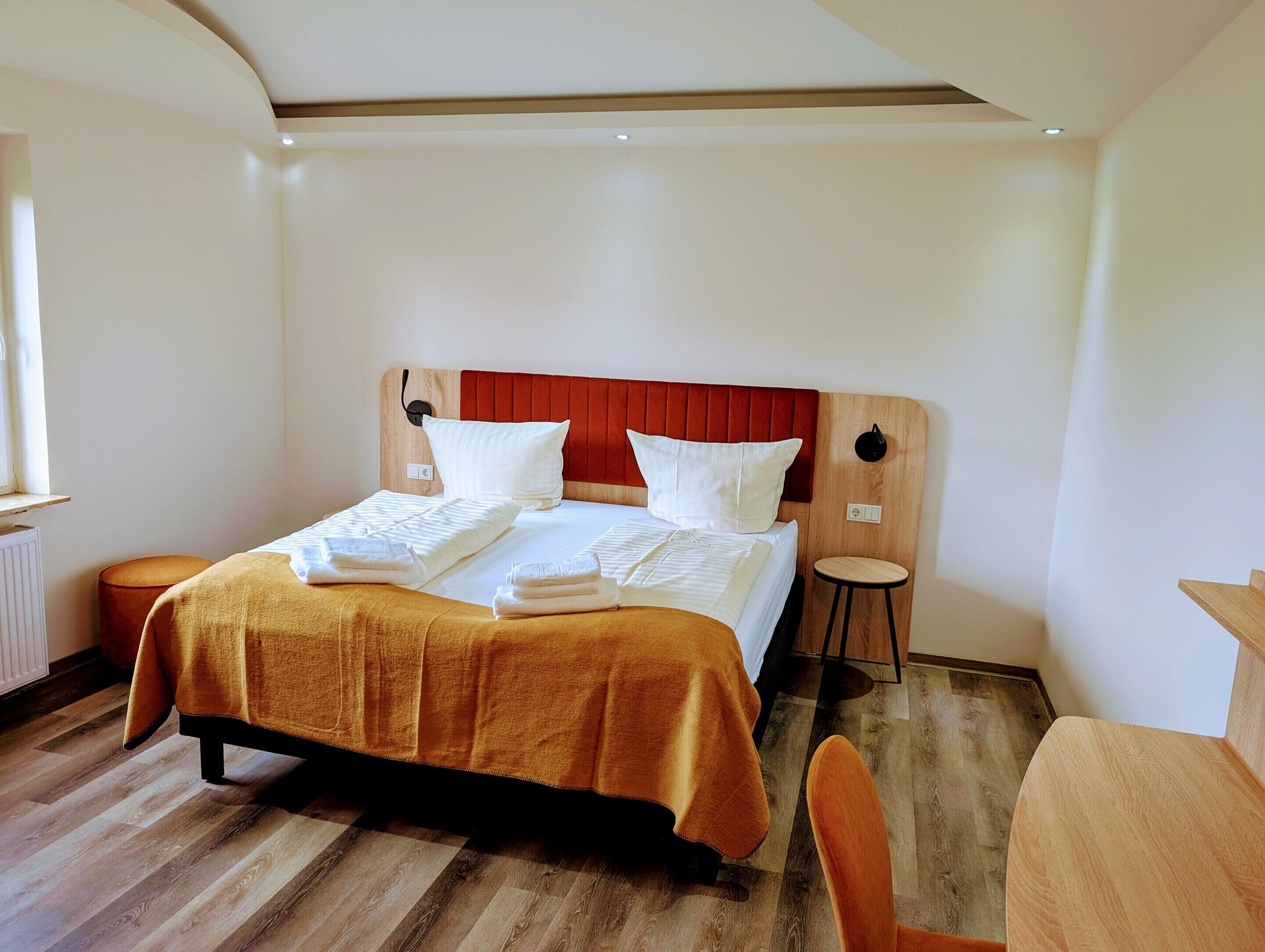 A hotel room with a bed, a wooden headboard, a small stool, and a wooden table.
