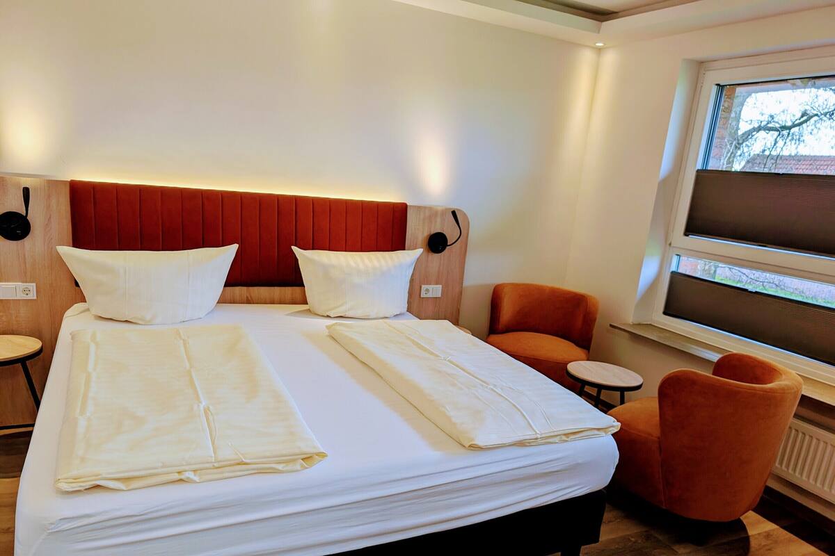 A hotel room with a large bed, two chairs, a small table, and a window with blinds.