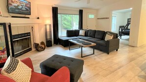 Una sala de estar con chimenea, sofá, reposapiés y televisor montado en la pared.