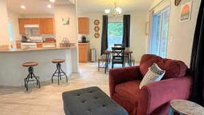 Una sala de estar con un sofá rojo, un reposapiés negro y una mesa de centro de madera. Hay una zona de cocina con armarios de madera y una barra.