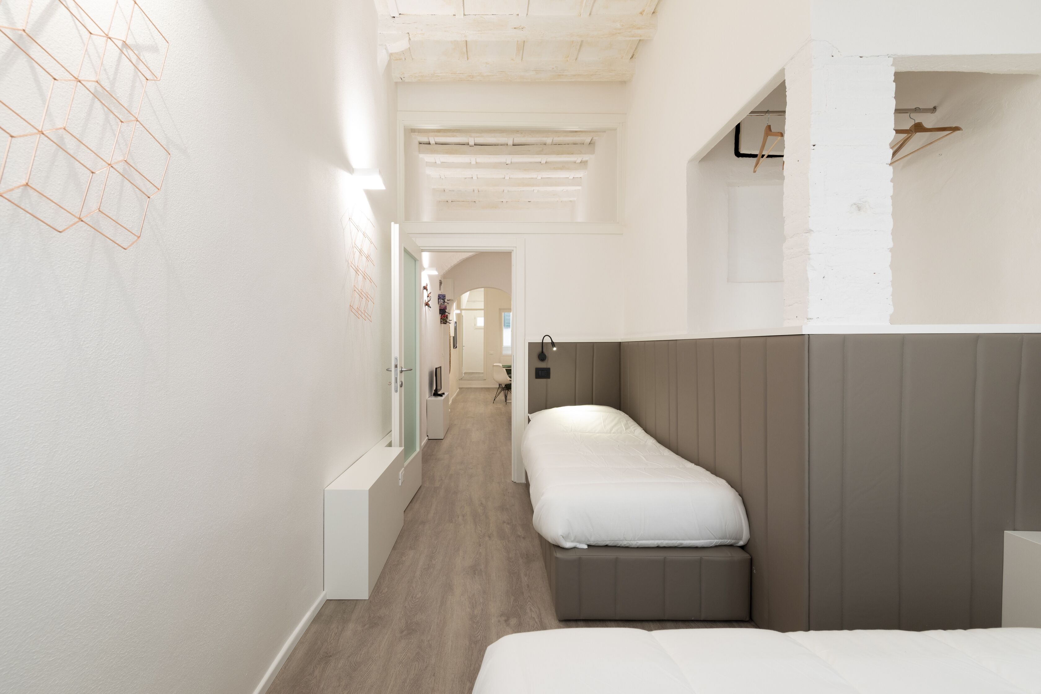 A narrow hallway with two single beds, a wooden floor, and a white wall with a geometric pattern.