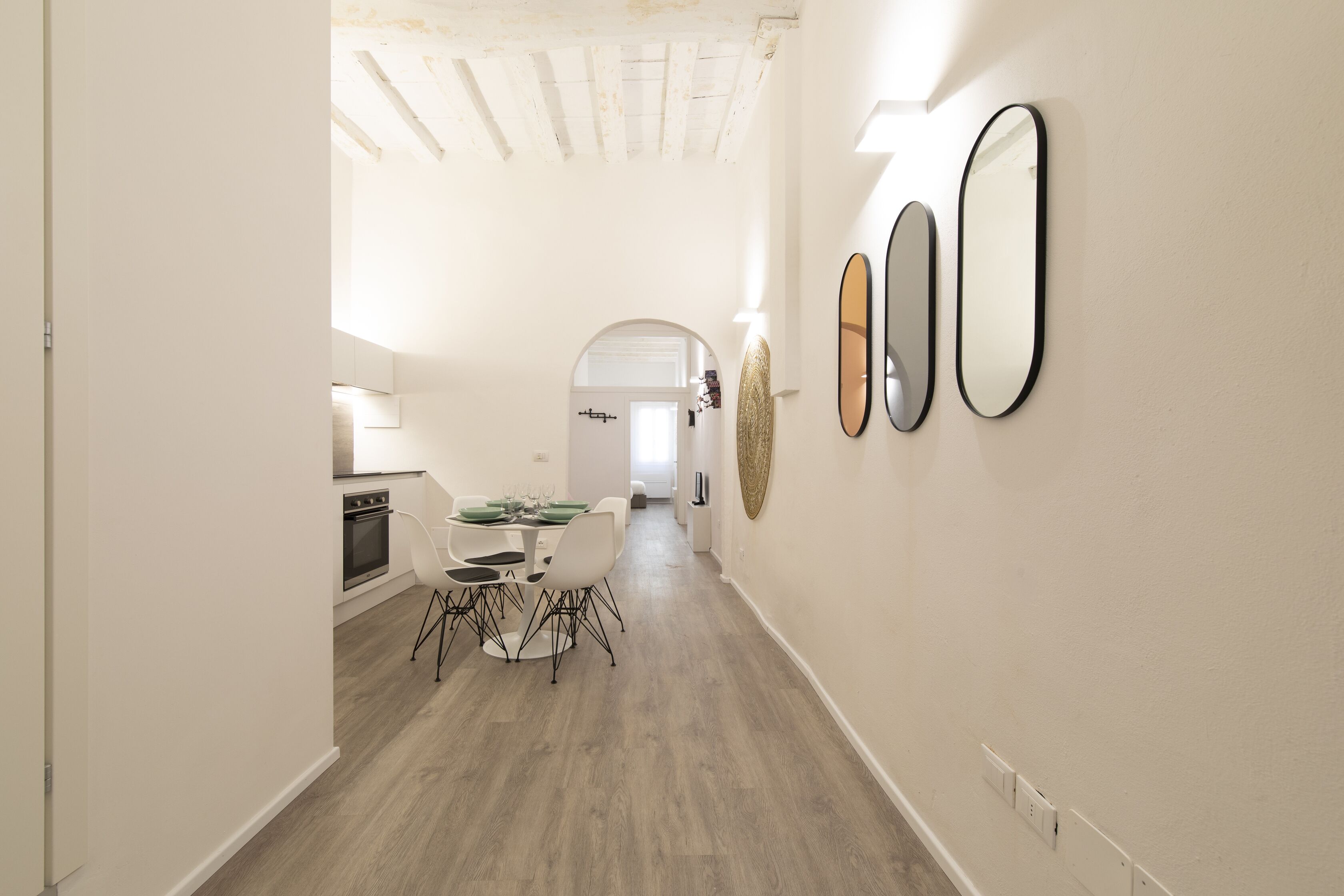 A narrow hallway with a dining area featuring a table and chairs, a kitchen with a stove, and a wall with three mirrors.