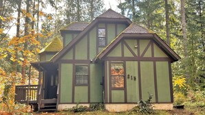 A green two-story house with a steep gabled roof, surrounded by trees and a wooden deck. - Mossy Moose (Packwood)