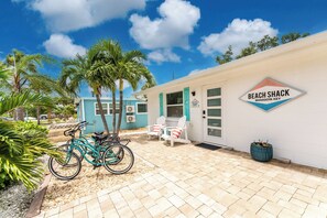 Uma cabana à beira-mar com uma placa indicando 'BEACH SHACK MANASOTA KEY', uma bicicleta estacionada na frente e um conjunto de mesa e cadeira para refeições ao ar livre.