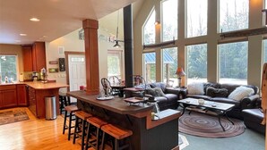 A spacious kitchen with a central island, bar stools, and a dining area with a sofa and coffee table. Large windows provide natural light and views of the outdoors. - Elkhorn (Packwood)